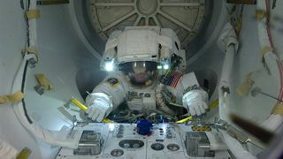 an astronaut inside an airlock grasping on to handles. in front is a panel with switches and dials
