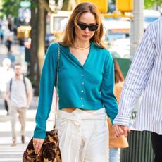 Jennifer Lawrence walks down the street in New York holding her husband's hand wearing white cotton trousers with a blue cardigan and beige chunky sandals.