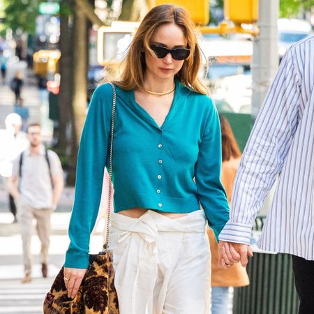 Jennifer Lawrence walks down the street in New York holding her husband's hand wearing white cotton trousers with a blue cardigan and beige chunky sandals.