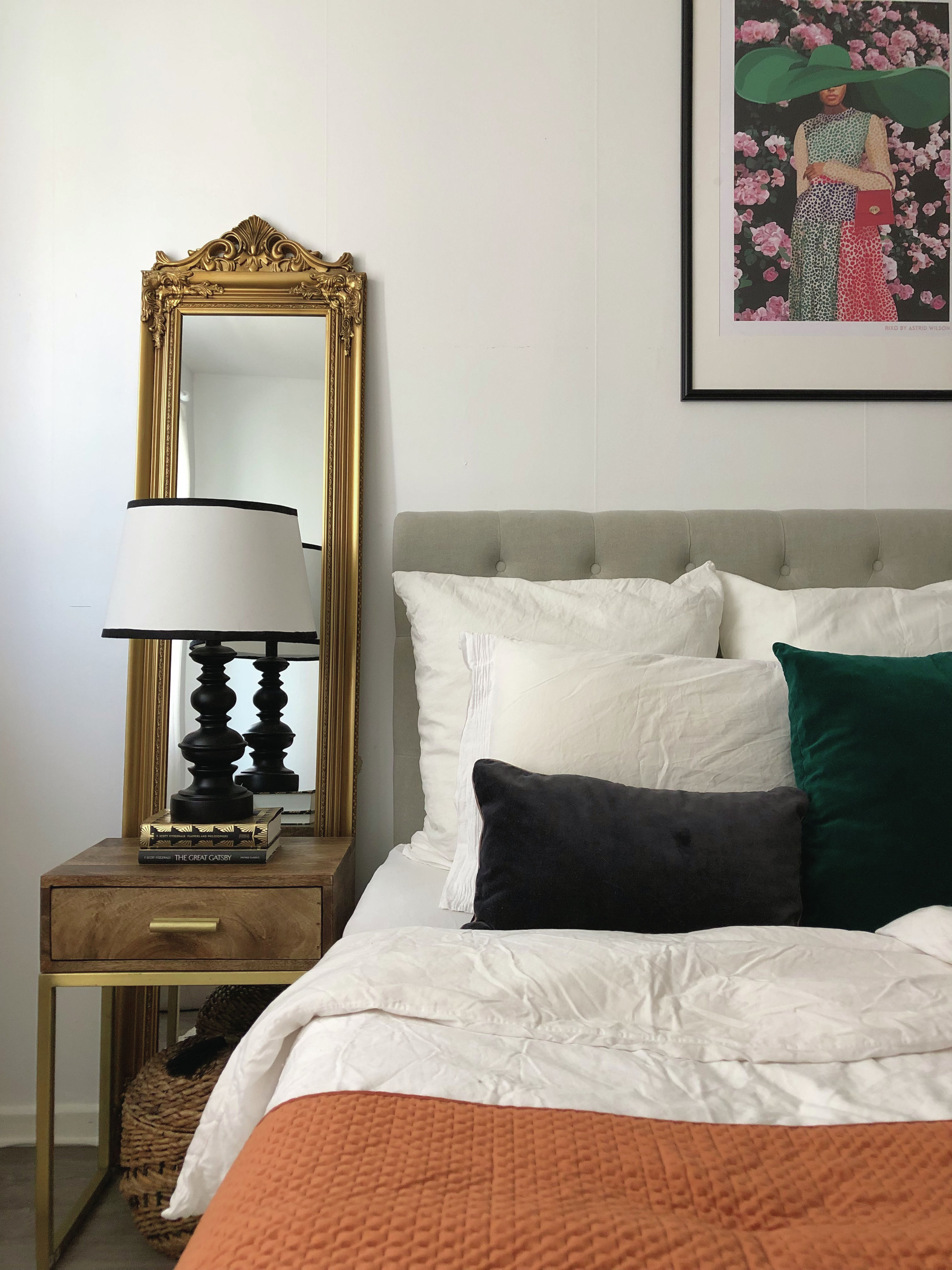A grey bedroom with gold mirror, cream and black lampshade and framed wall art of woman