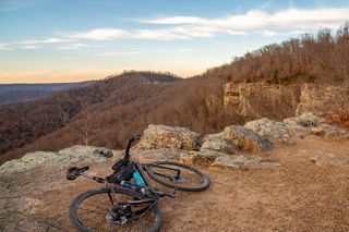 Una bicicleta gravel negra yace de lado en la tierra color canela de Arkansas, con vistas a las montañas de Boston, que son escasas y aún no tienen hojas.