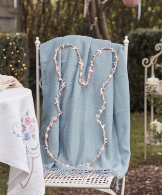 A white iron chair outdoors with a blue throw draped over the top of it and a bunny shaped LED wreath on it, with a white floral tablecloth to the left of it and an ornate garden shelf, a tree, and hedges behind it