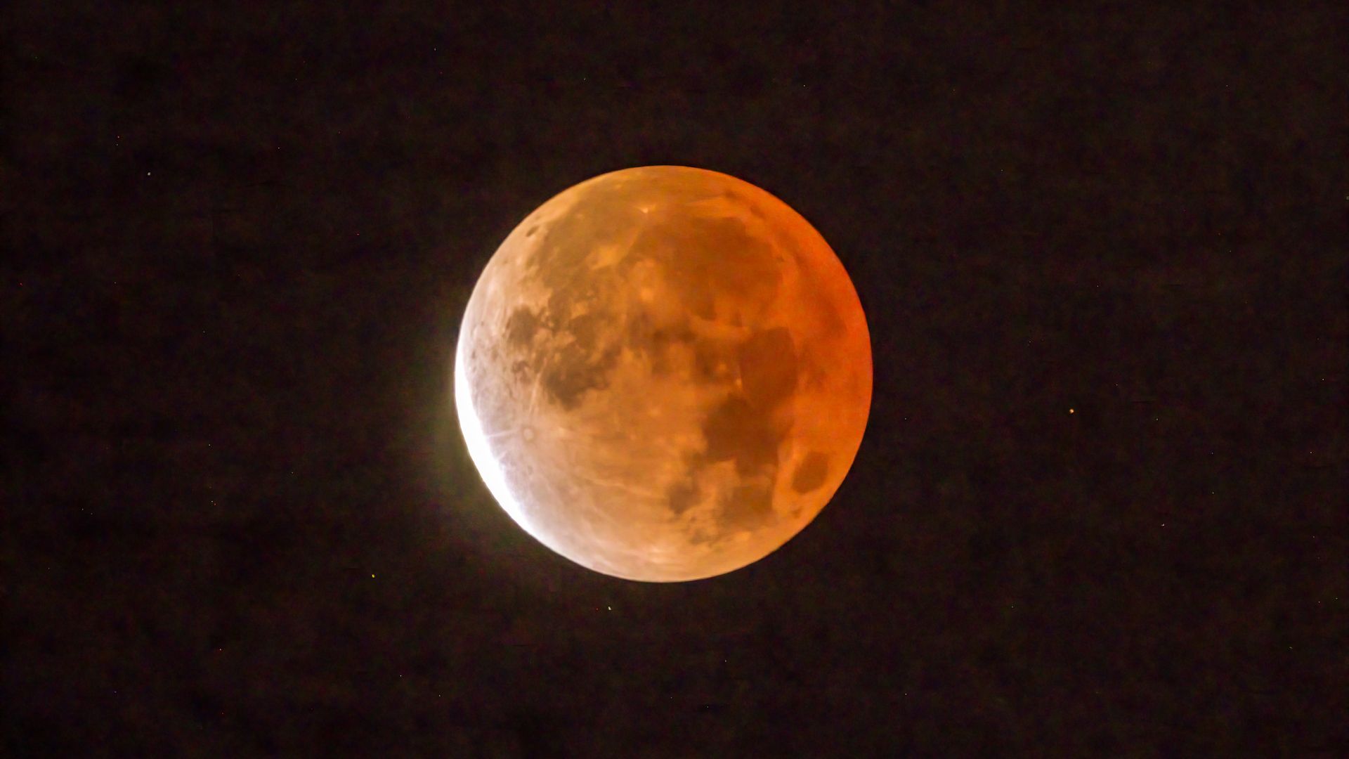 An orange-red moon is pictured during a lunar eclipse in a black sky. Craters and dark lunar seas are visible on its surface and a bright crescent shines on the left where it is exposed to sunlight