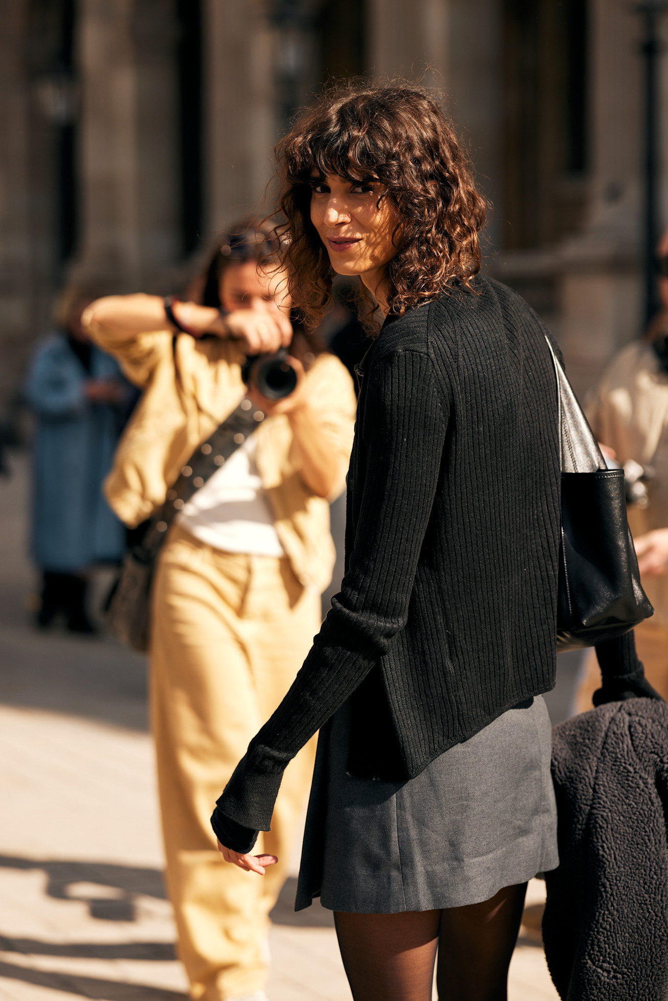 a woman with curly bangs and a curly lob wearing a black sweater and gray skirt looking over her shoulder