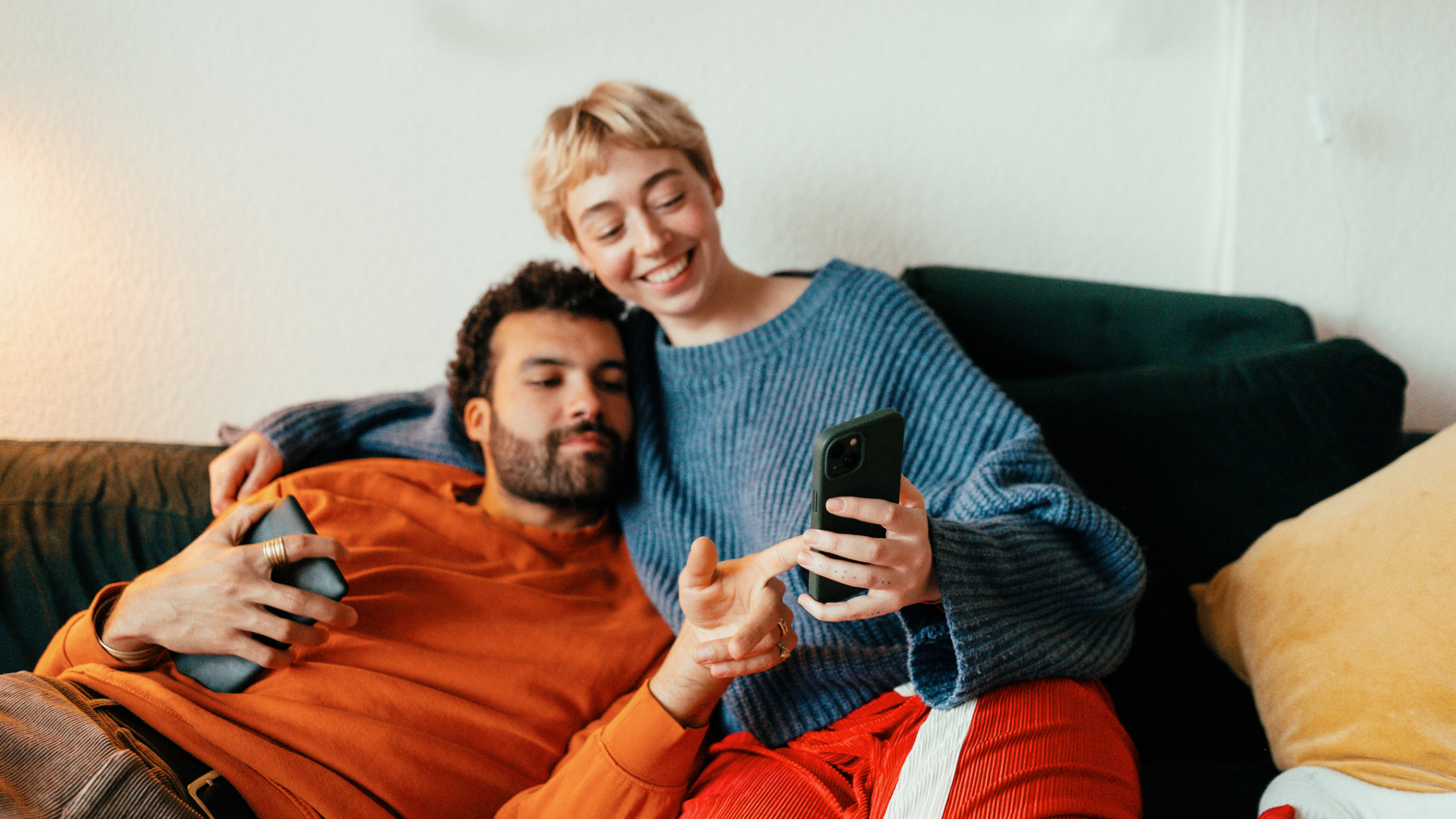 Two people, sat on a sofa, holding their phones and pointing at one of them.