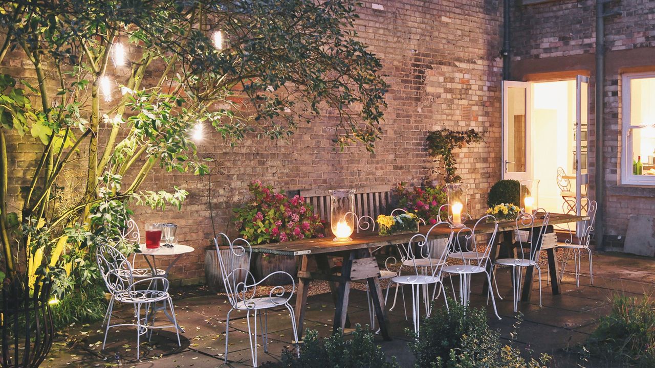 Garden patio at nighttime, featuring a long wooden dining table, metal chairs, and lighting throughout