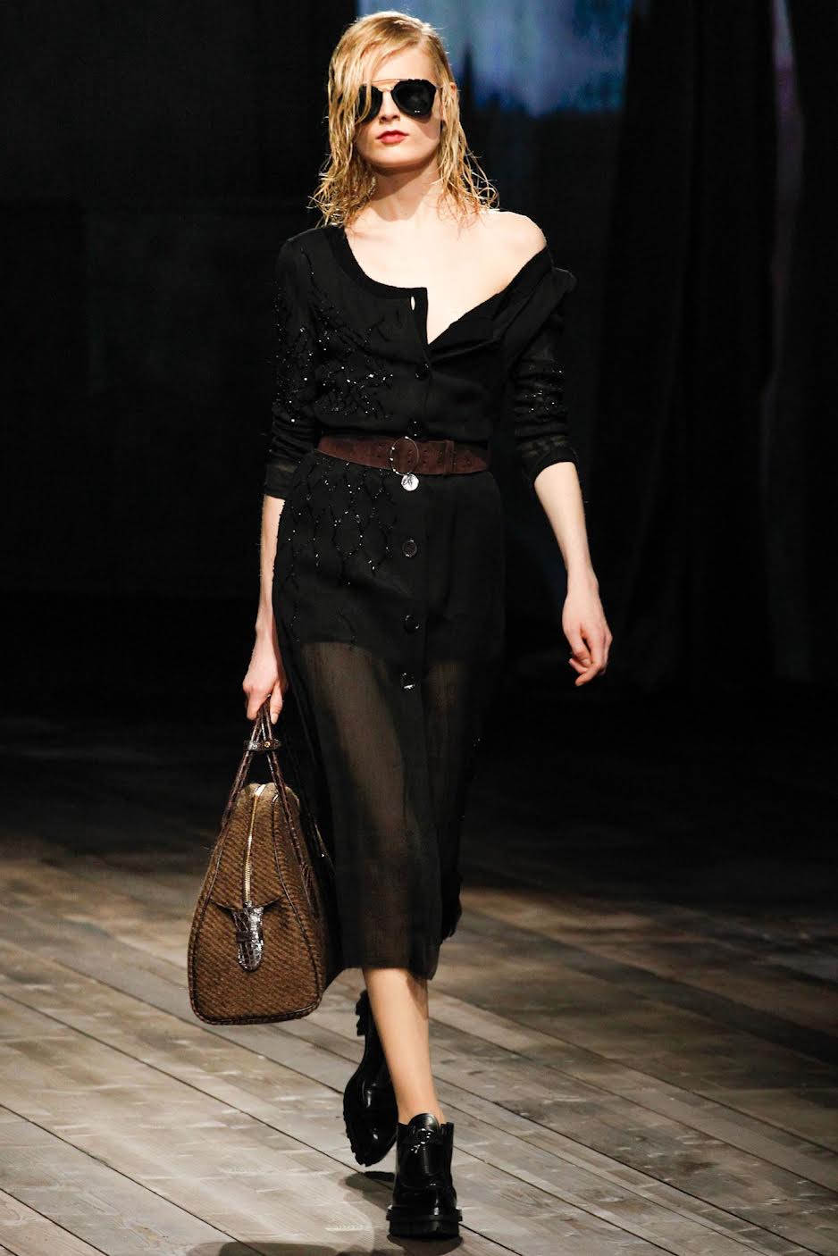 a model walking in Prada 2013 wearing a thin black knit cardigan and a sheer skirt with a brown belt and bag