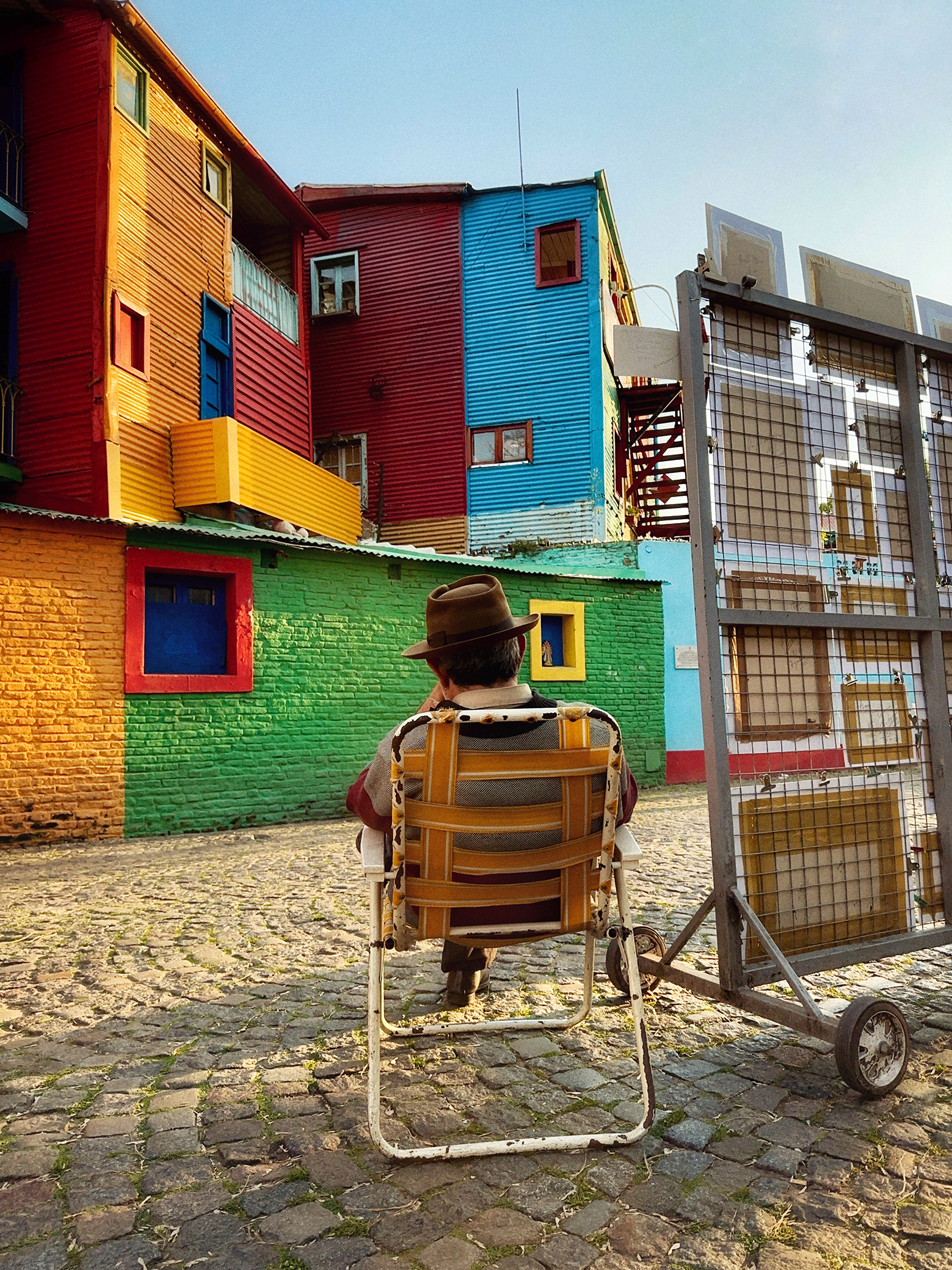 An elderly man in a hat sits in a folding chair, gazing at vibrant, multi-colored buildings in a cobblestone street