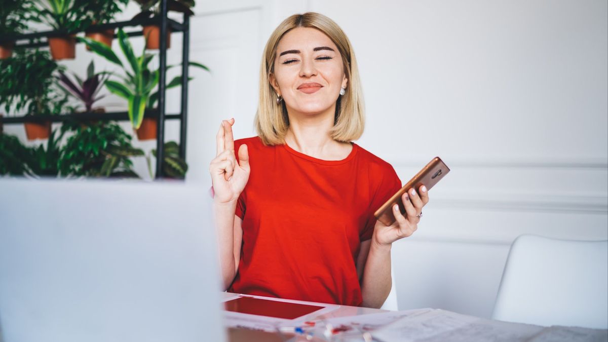 young woman with dyed blonde hair and a bright red shirt sits in front of a laptop; her eyes are closed and her fingers are crossed, as if she&#039;s making a wish. In one hand, she holds a cell phone.