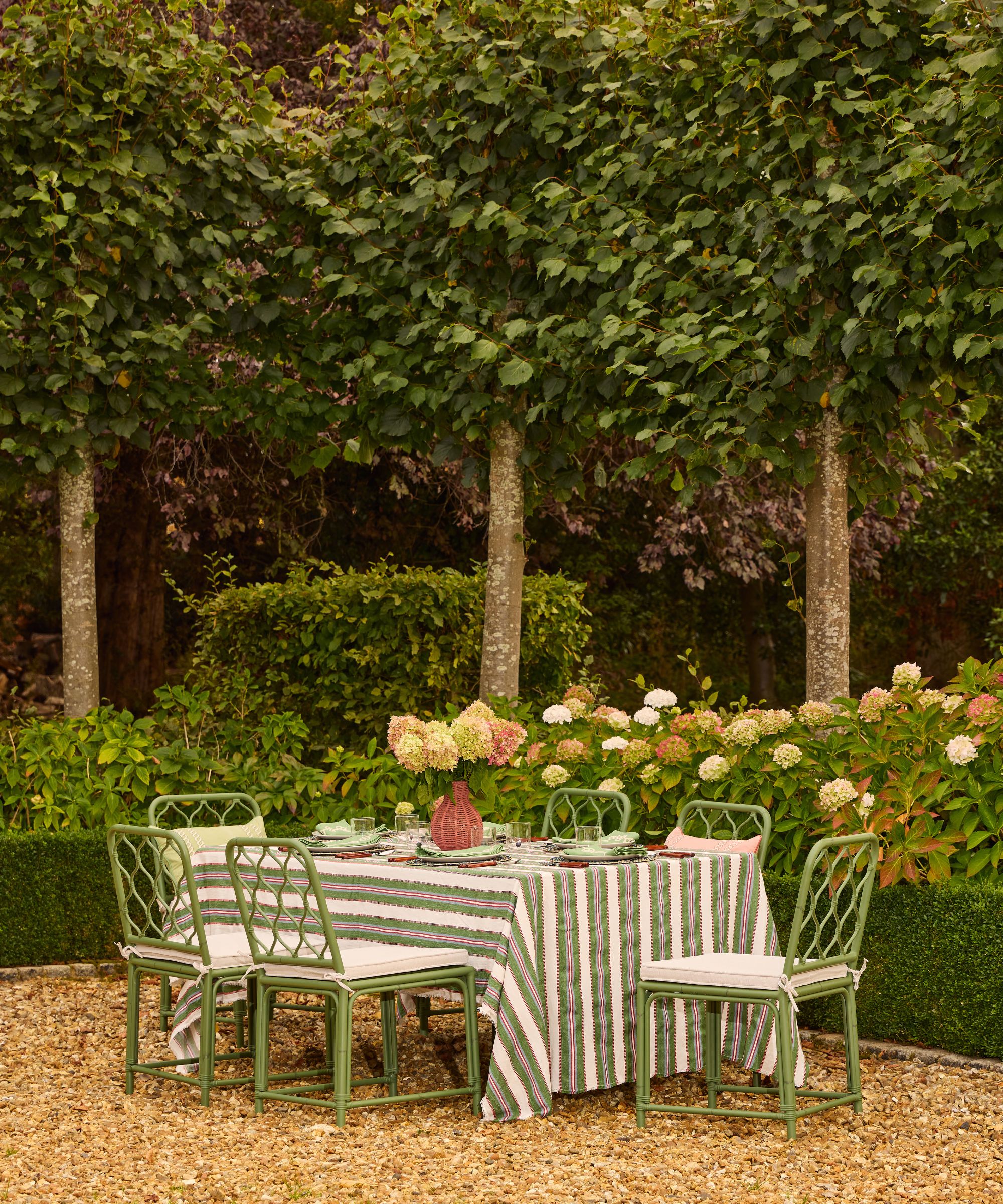 a landscape garden with tall hedges and trees, a border of hydrangeas, and a shingle patio with green bamboo garden dining furniture with a striped tablecloth