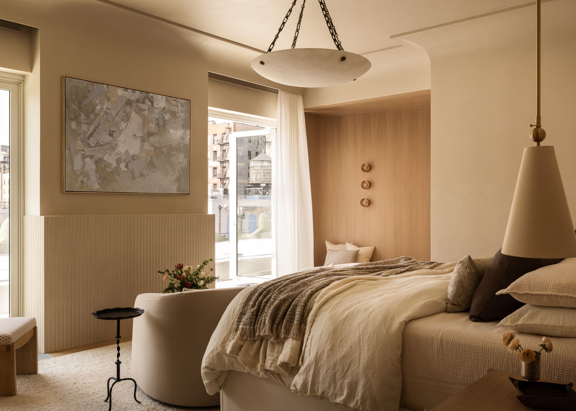 A neutral modern bedroom with cream walls, pendant lighting, a piece of art hangs between the two windows facing the bed which has armchairs in front of it