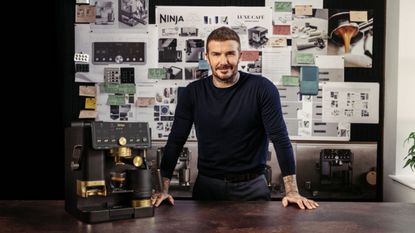 Image of David Beckham standing in front of a bulletin board with papers pinned on it in a long-sleeve navy shirt with a gray and gold espresso machine next to him.