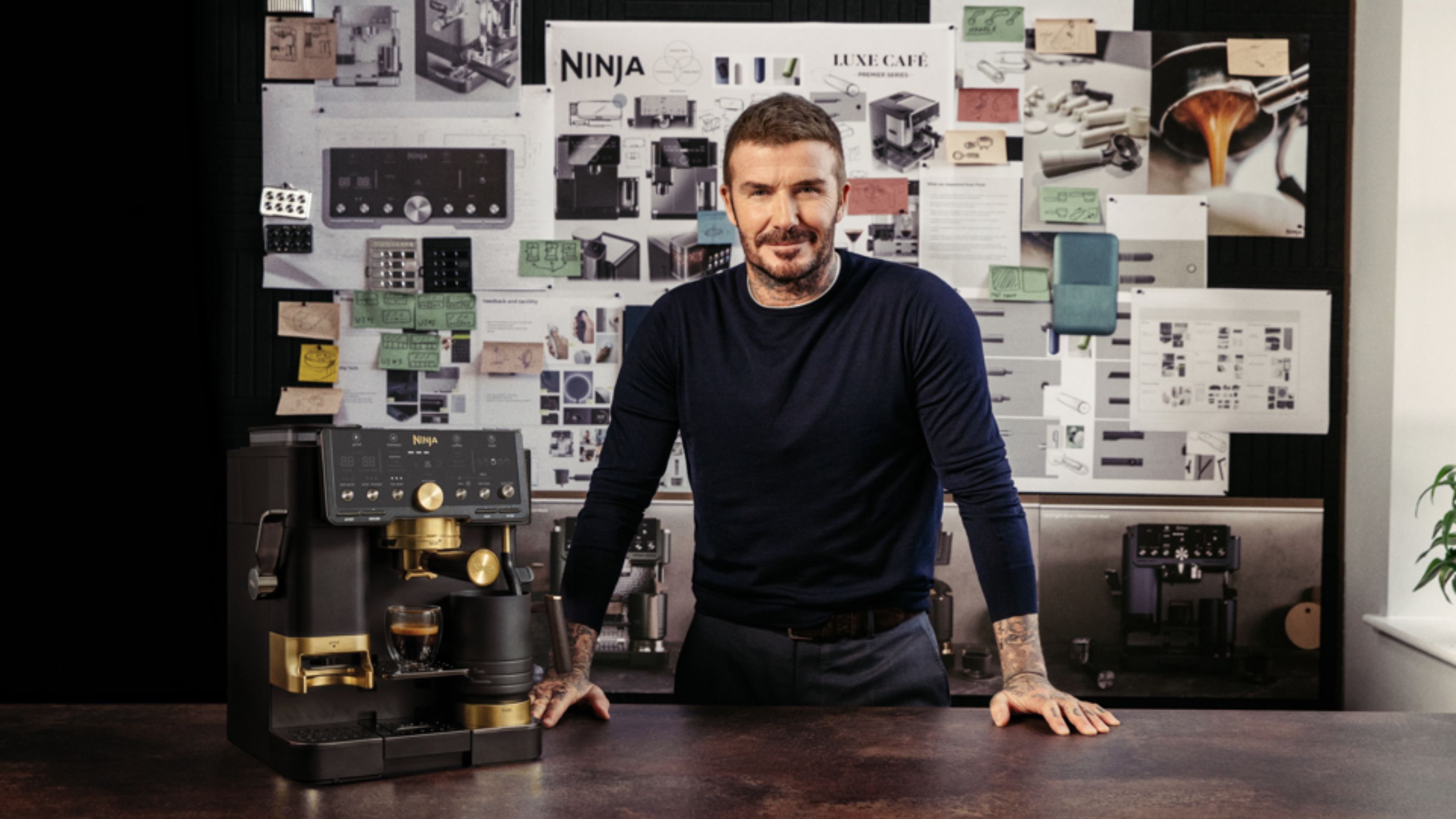 Image of David Beckham standing in front of a bulletin board with papers pinned on it in a long-sleeve navy shirt with a gray and gold espresso machine next to him. 