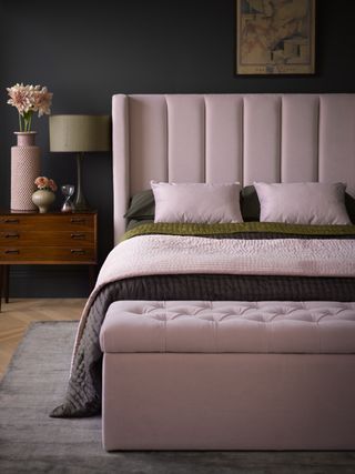stunning pink and green bed with pink ottoman, wooden bedside table with accessories on top, a light wooden floor and a thin grey rug, against a dark black wall
