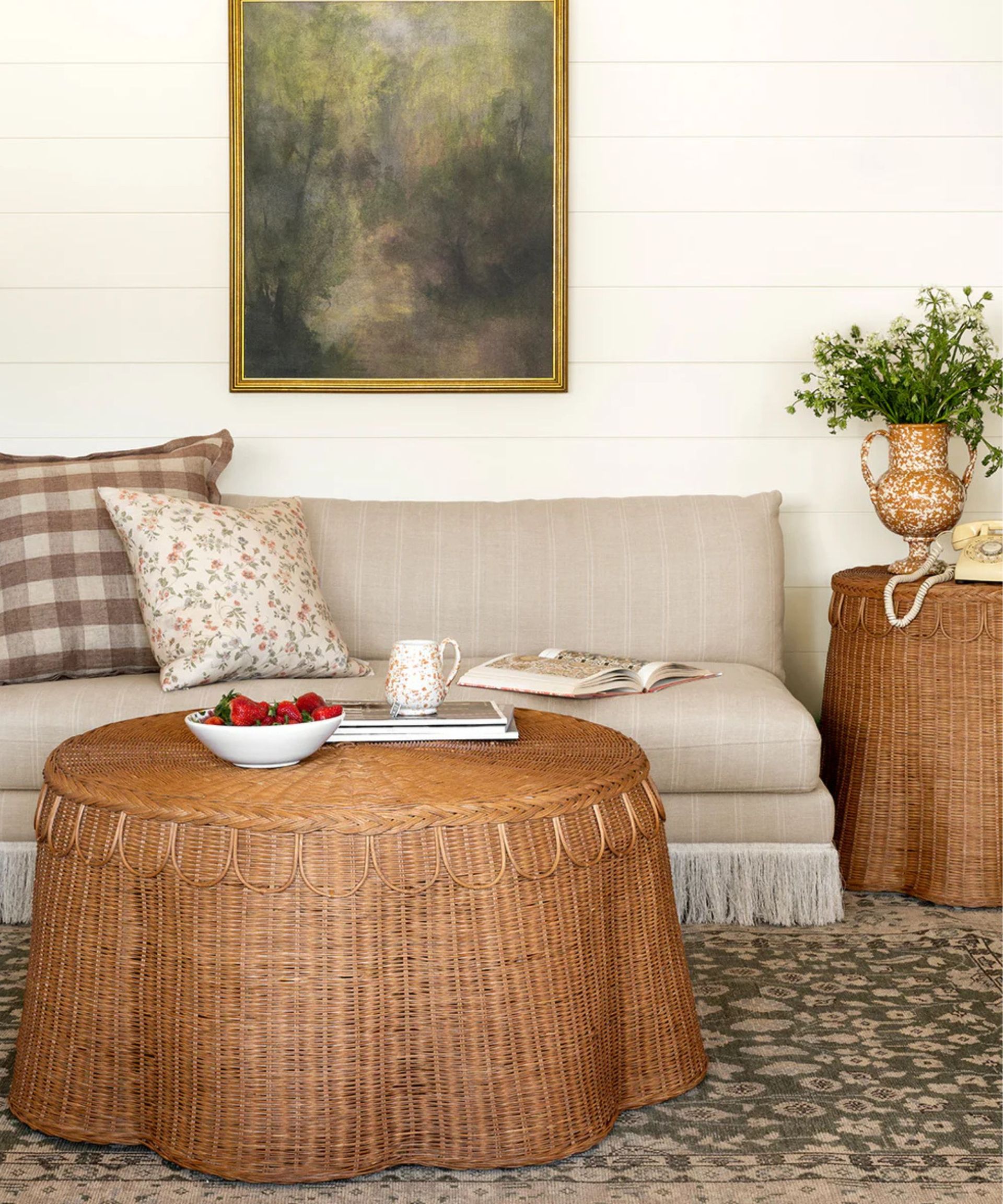 mgee and co summer living room spot with a striped beige armless sofa with fringed trim style with a plaid and floral cushions and a rattan scalloped round coffee table and matching side table styled with splatter vase with florals