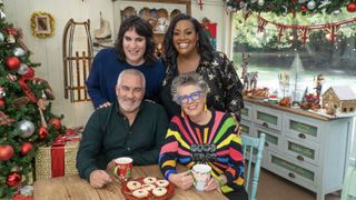 Paul Hollywood in a dark shirt sits with Dame Prue Leith in a rainbow jumper and Noel Fielding in a dark jumper and Alison Hammond in a starry blouse stand behind them in the tent which is decorated for Christmas in The Great British Bake Off.