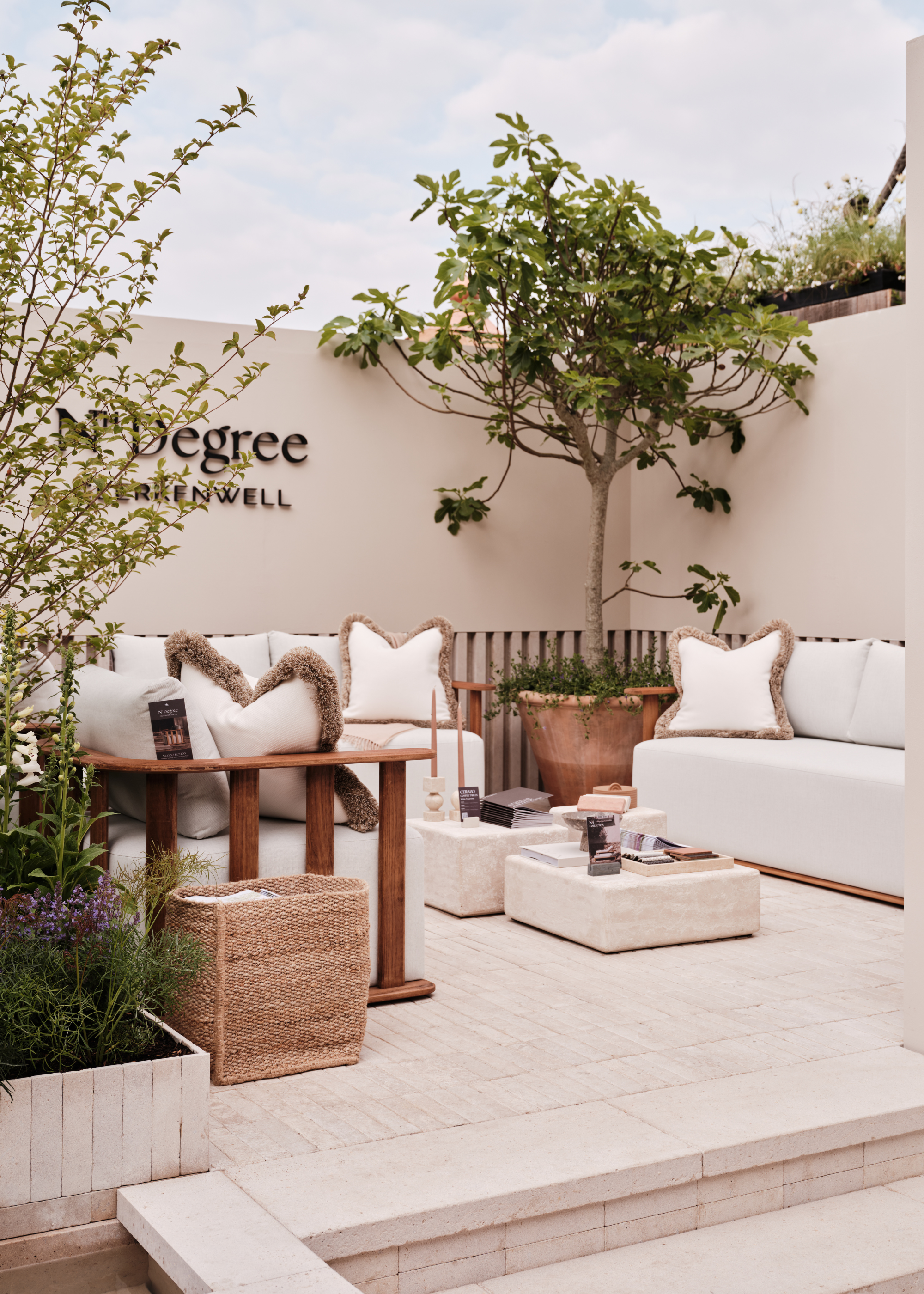 A still from Nth Degree's exhibit at the 2025 Chelsea Flower Show featuring outdoor sofas, block coffee tables, a woven basket, a shrub in a trough-style container, and a potted tree