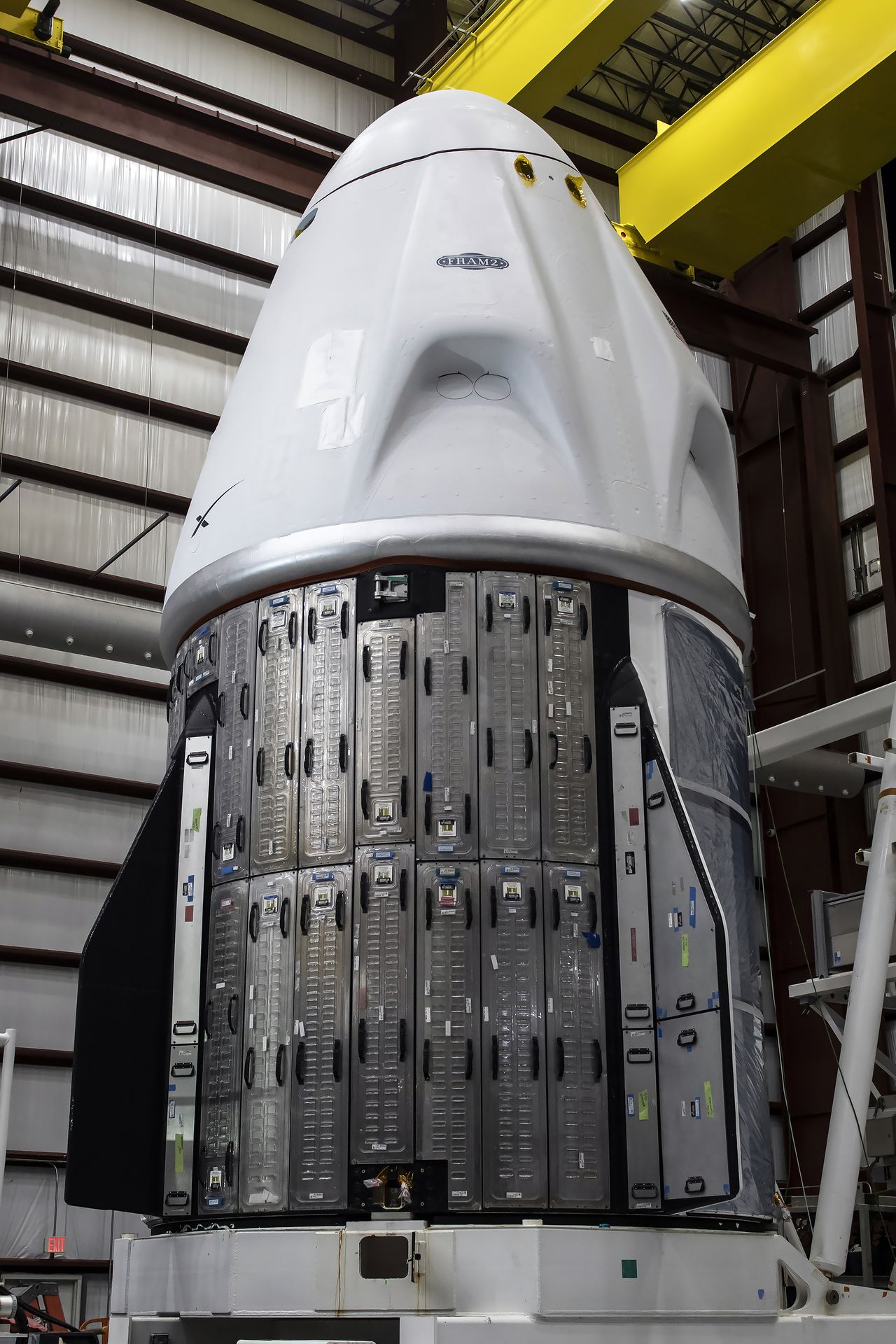 A white conical space capsule stands atop a metal trunk support inside a hangar.