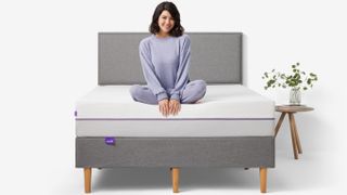 Purple Original vs Purple Plus Mattress: A woman with dark hair and dressed in lilac coloured pyjamas sits on the edge of the Purple Plus Mattress Purple Plus Mattress
