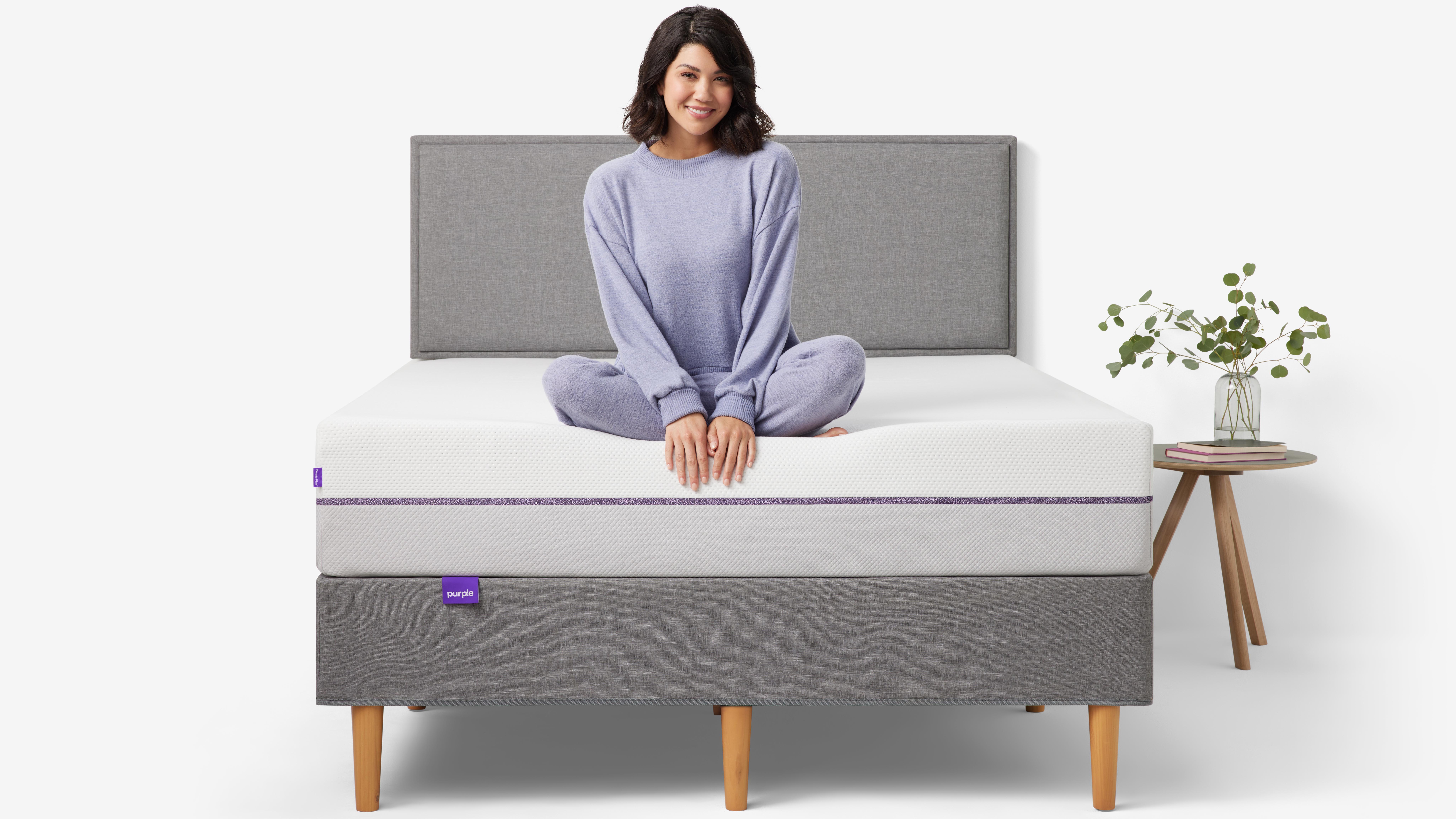 Best mattress: A woman with dark hair and dressed in lilac coloured pyjamas sits on the edge of the Purple Plus Mattress