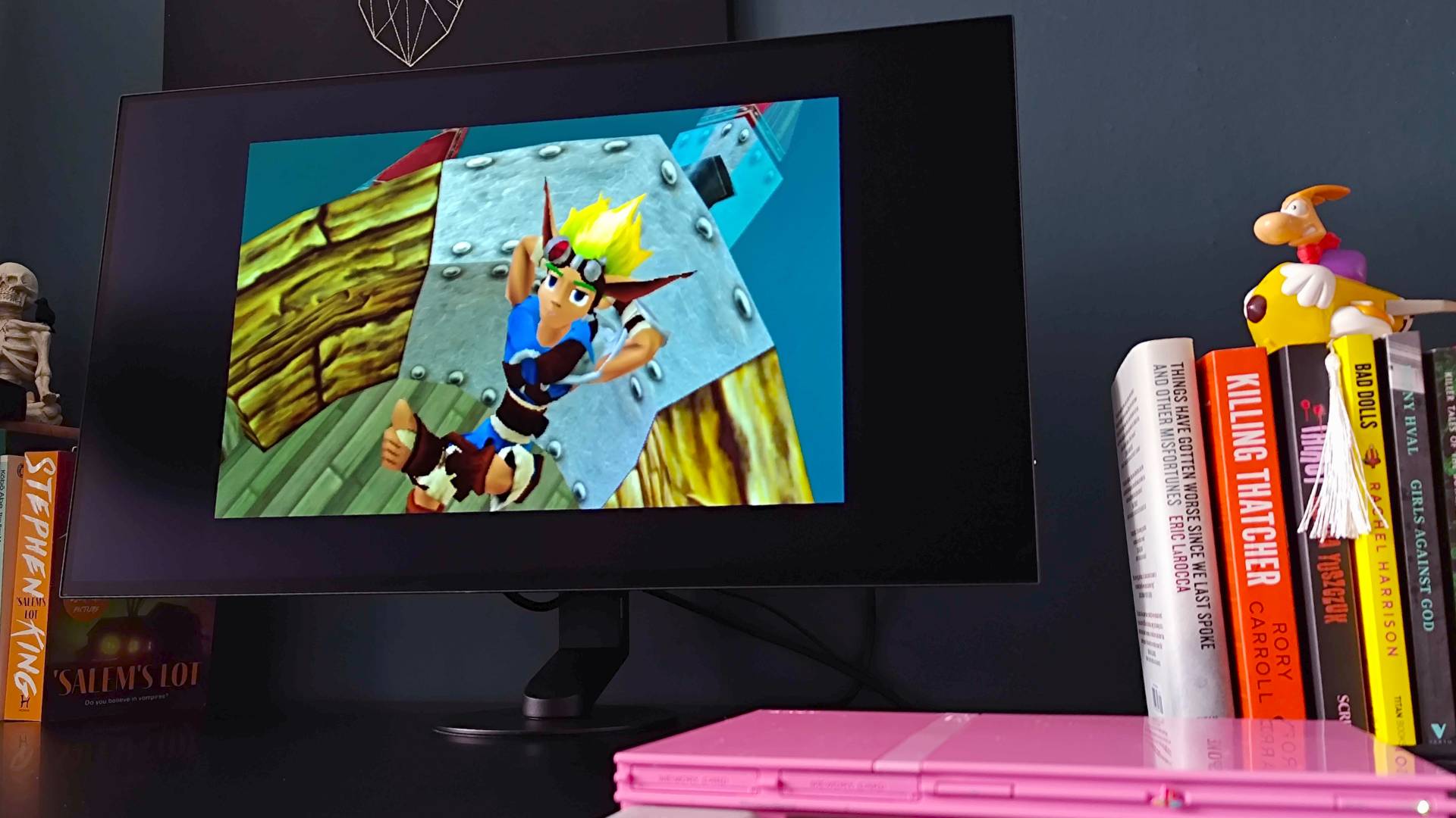 Sony Inzone M10S II on black desk next to books and pink PS2 slim with Jak and Daxter cutscene on display.