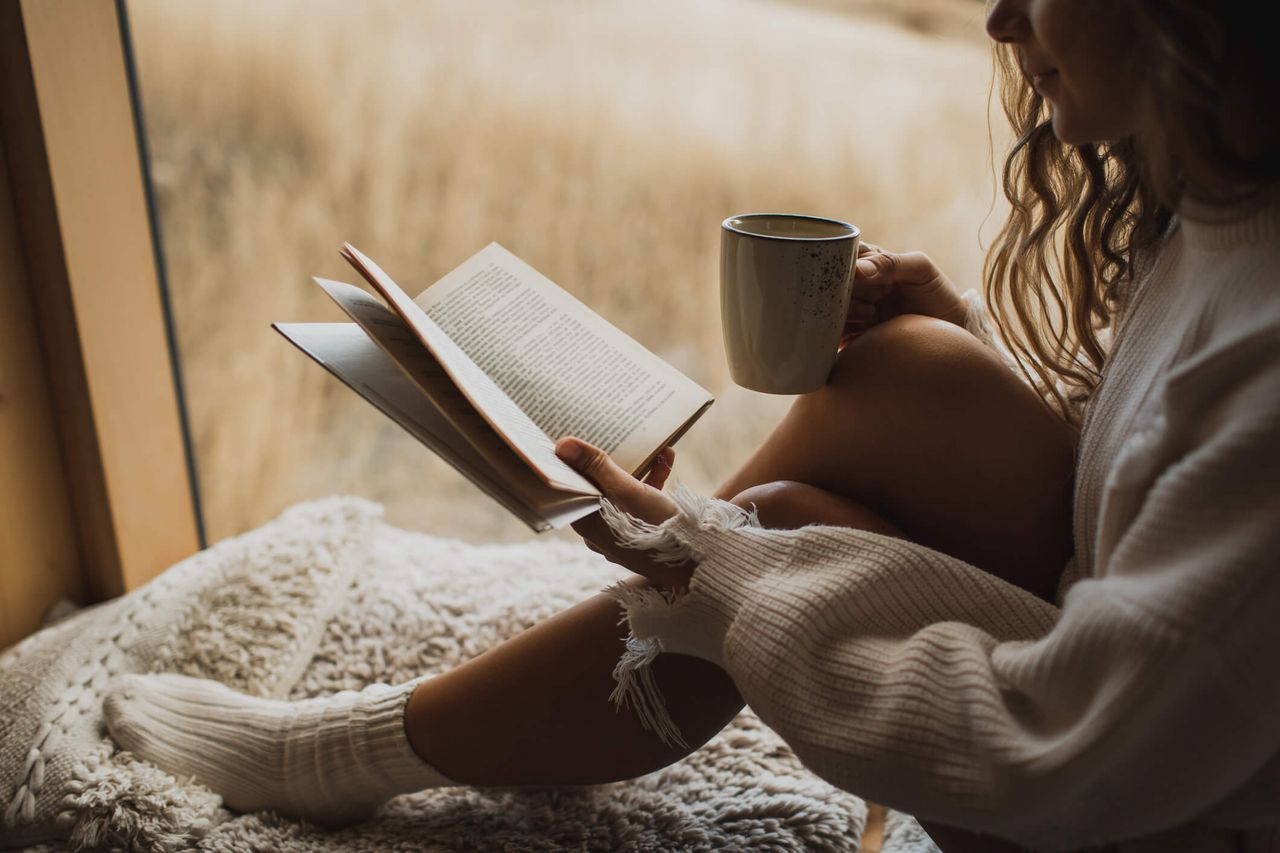 A woman reading a book and enjoying a warm drink for a blog article on autumn activities.