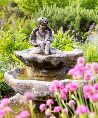 A silver tiered garden sculpture with a cherub figure on top, pink tall flowers in front of it, and a large lime green hedge behind it