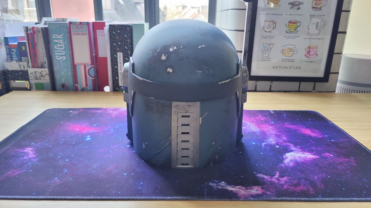 The Star Wars: Black Series Bo-Katan Helmet with the heads-up display up. There is a gray, black, and white owl-like pattern on the front of the light blue helmet and a black T-shaped visor. This photo shows the back of the helmet.