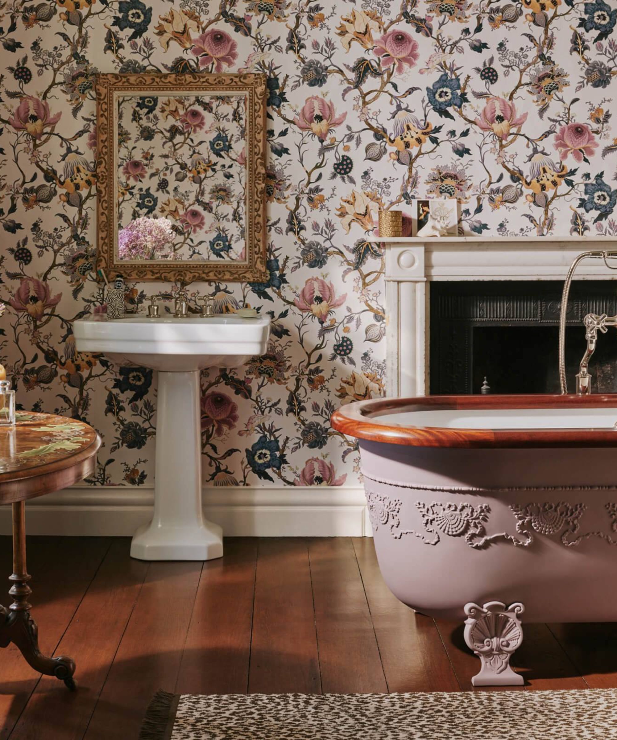 A bathroom with floor wallpaper, a white pedestal sink, and a pink freestanding bath with floral carvings