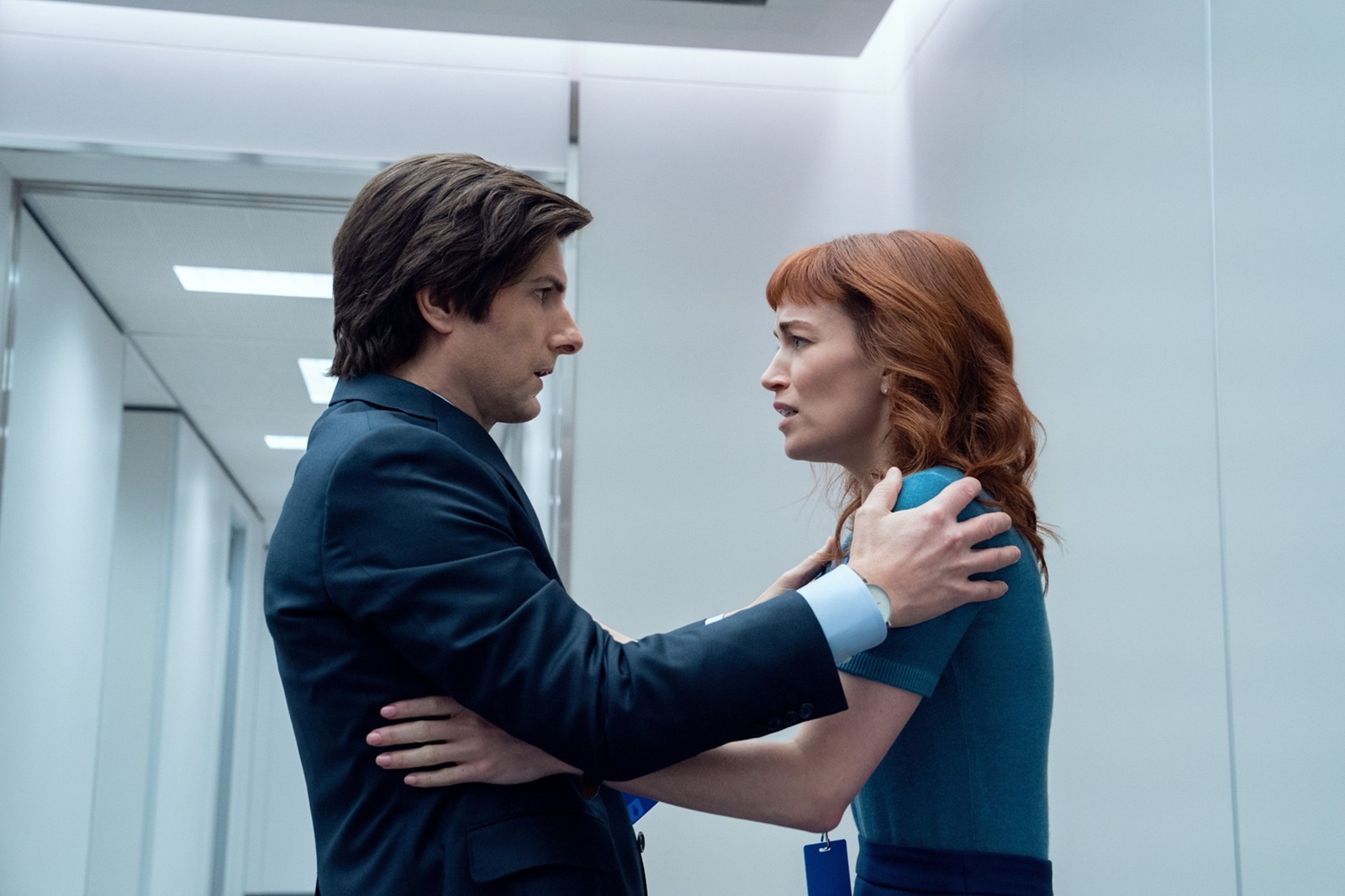 A man and a woman in office attire stand in a stark white hallway, holding onto each other's shoulders and exchanging an intense, emotional look.