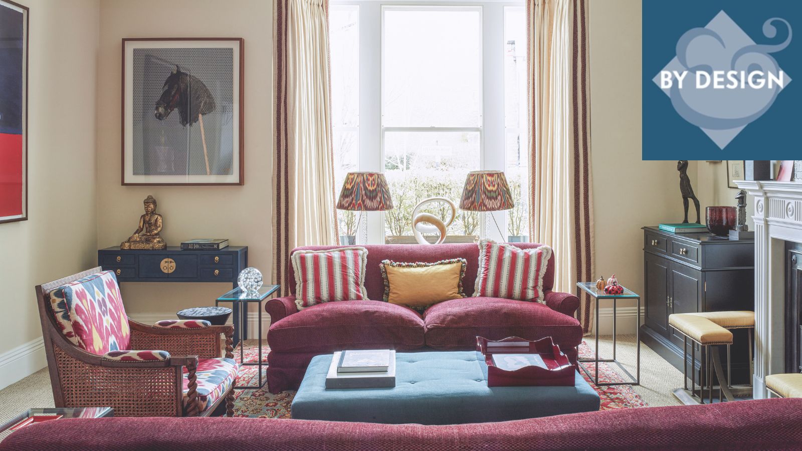 Living room with light yellow painted walls, cherry sofa, lounge chair and blue ottoman, artwork and lampshades in background near bay window