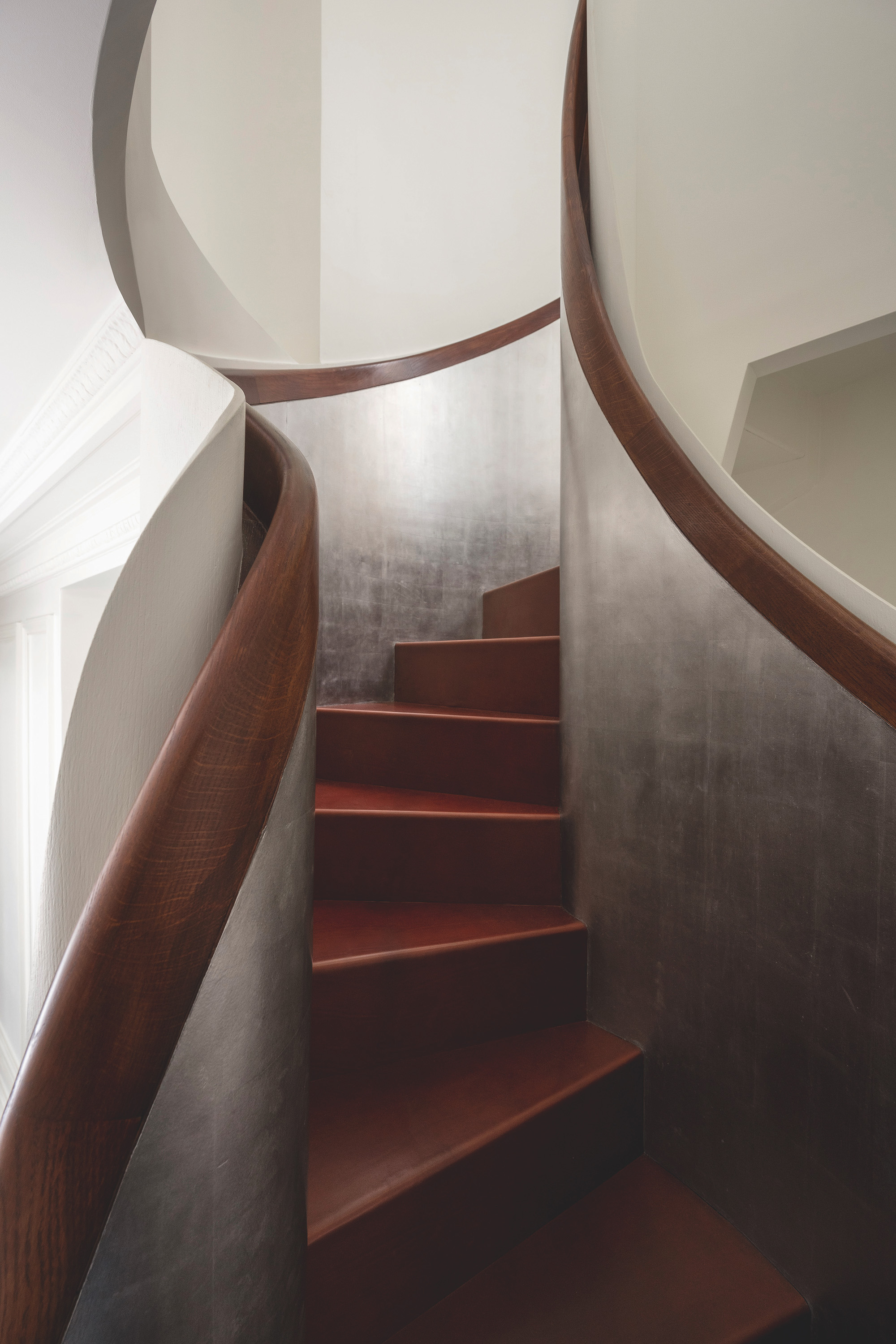 A curved staircase with with cream walls, dark wood handrails that transition in to a silver colour and red stairs.