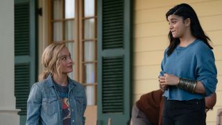Still from The Marvels (2023) movie. Carol and Kamala have a chat on the porch. Carol is looking up at Kamala who is standing on the porch outside a house. Carol is a woman with long blonde hair tied back in a low ponytail and is wearing a t-shirt and a denim shirt overtop. Kamala is a teen with long dark hair and is wearing a blue sweatshirt with the cuffed pulled up which reveals the magical bangle she is wearing on her forearm.
