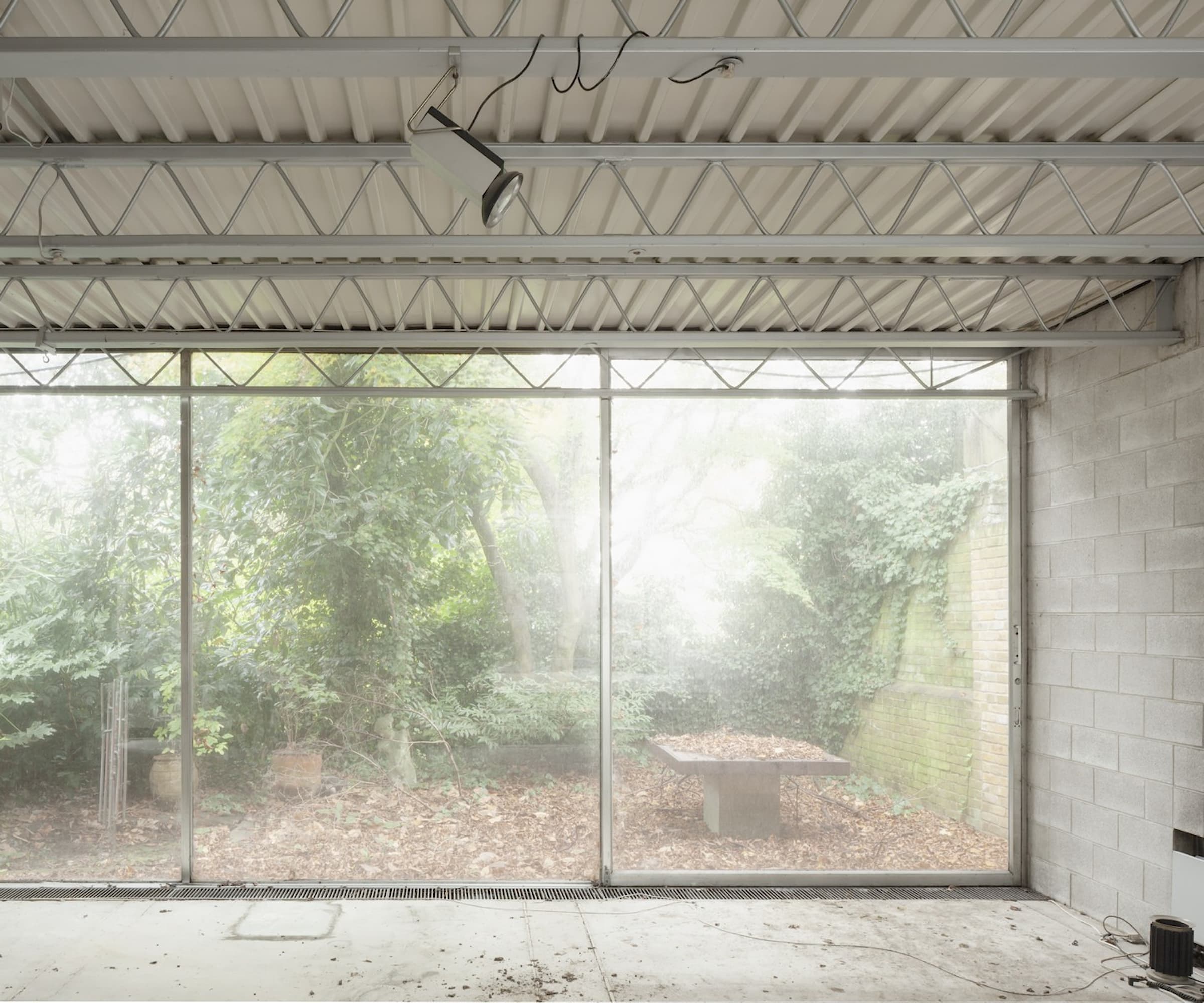 A living room with concrete walls and a floor to ceiling window looking onto an overgrown garden