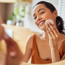 Best toner: Portrait of young woman looking at mirror while cleaning her face with cotton disc in bathroom. 