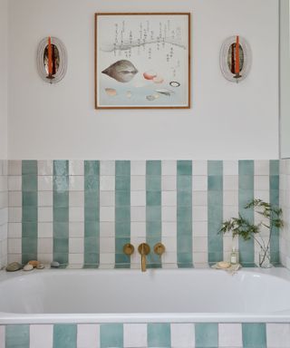 coastal bathroom with striped tiles above a deep white tub gold hardware and fish art work on wall