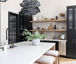 Modern kitchen space with white island and bar stools, beneath large statement ceiling pendant with open wooden shelving in the background