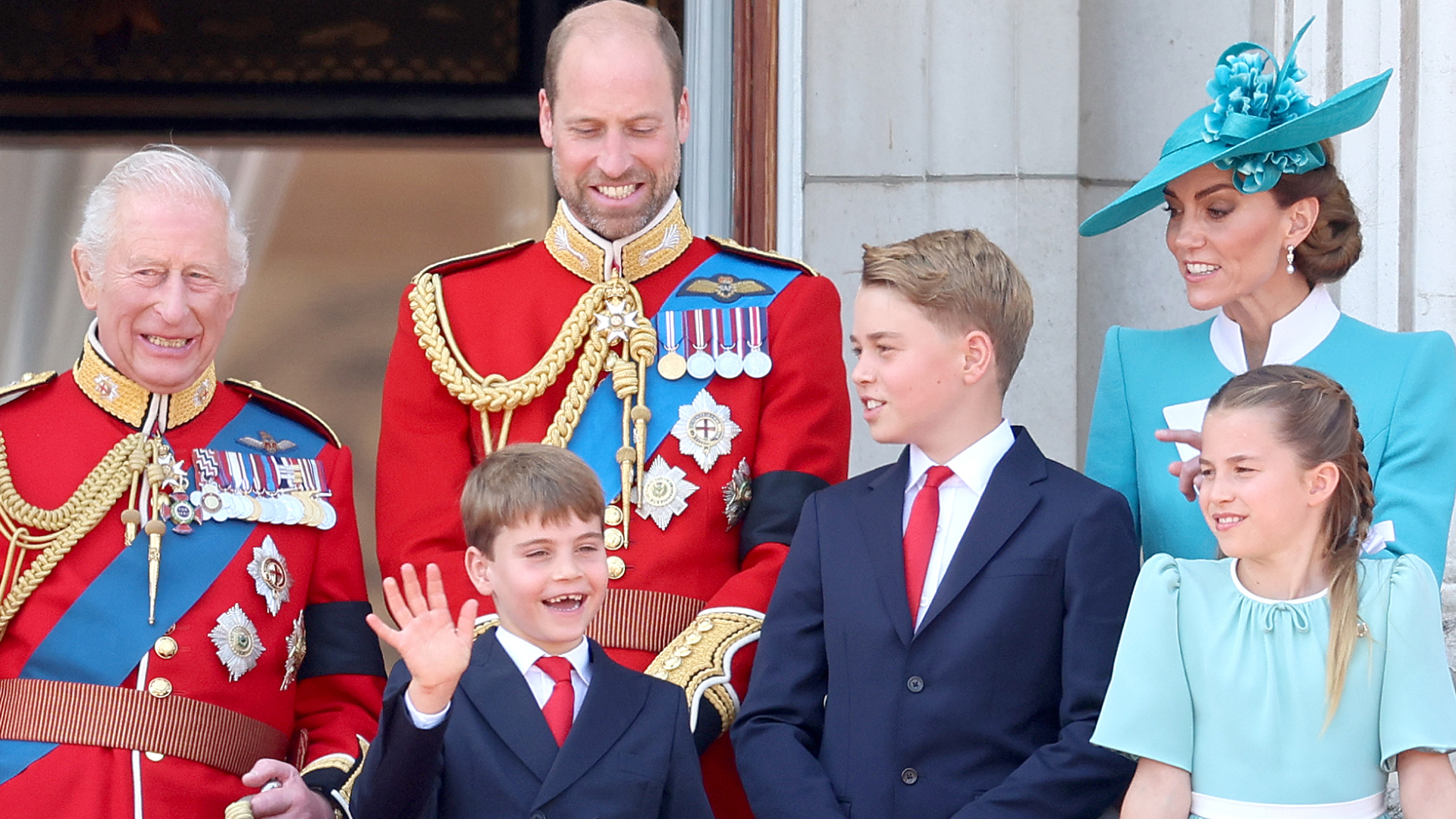 King Charles and Prince William wear red military uniform, Prince Louis and Prince George wear navy suits with red ties, while Princess Kate and Prince Charlotte wear turquoise dresses on the Buckingham Palace balcony