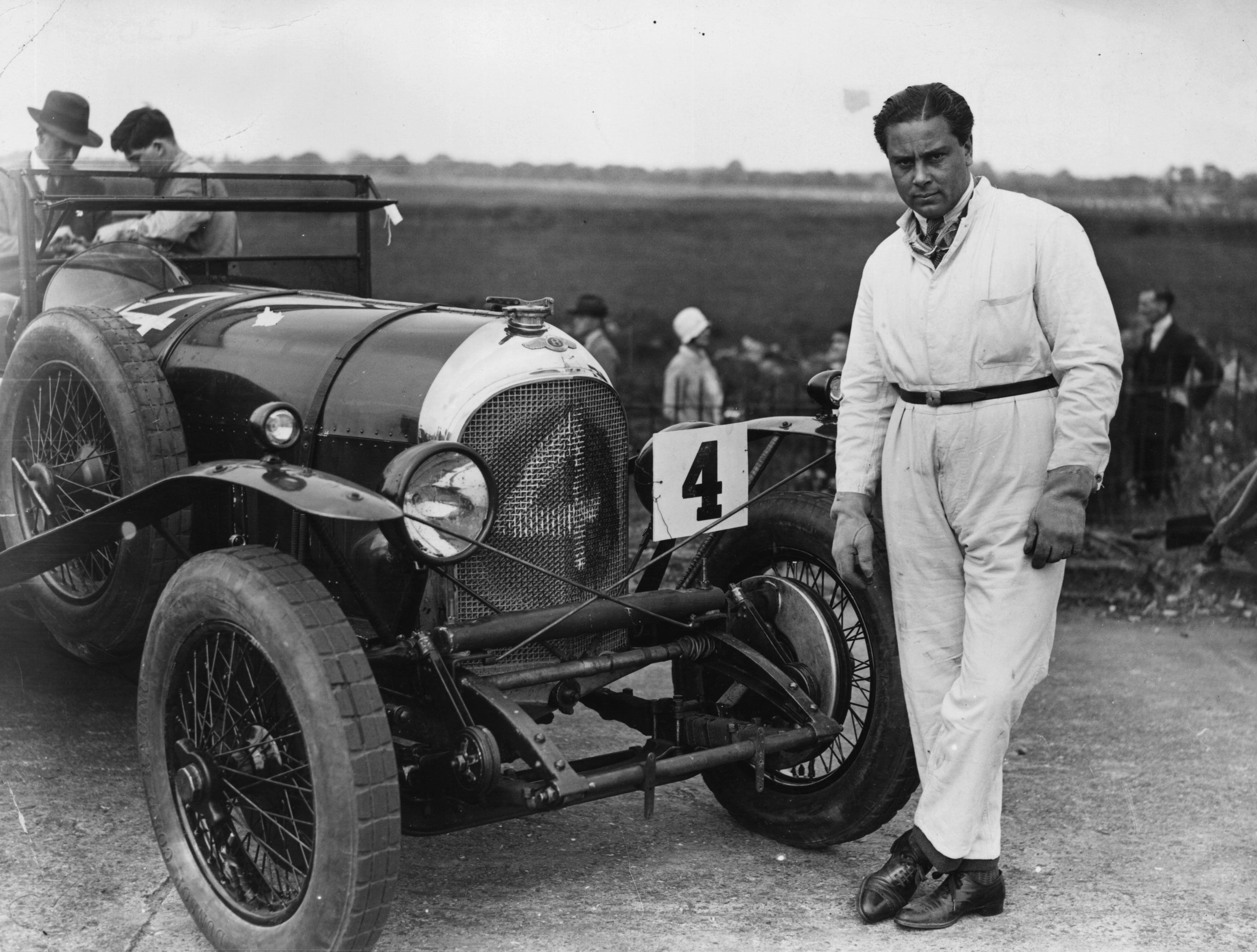A man in a white race suit standing next to a vintage Bentley
