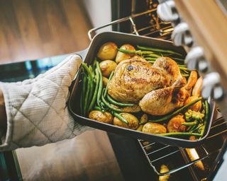 Roasted chicken and vegetables in a pan being pulled out of the oven from someone in beige oven mitts