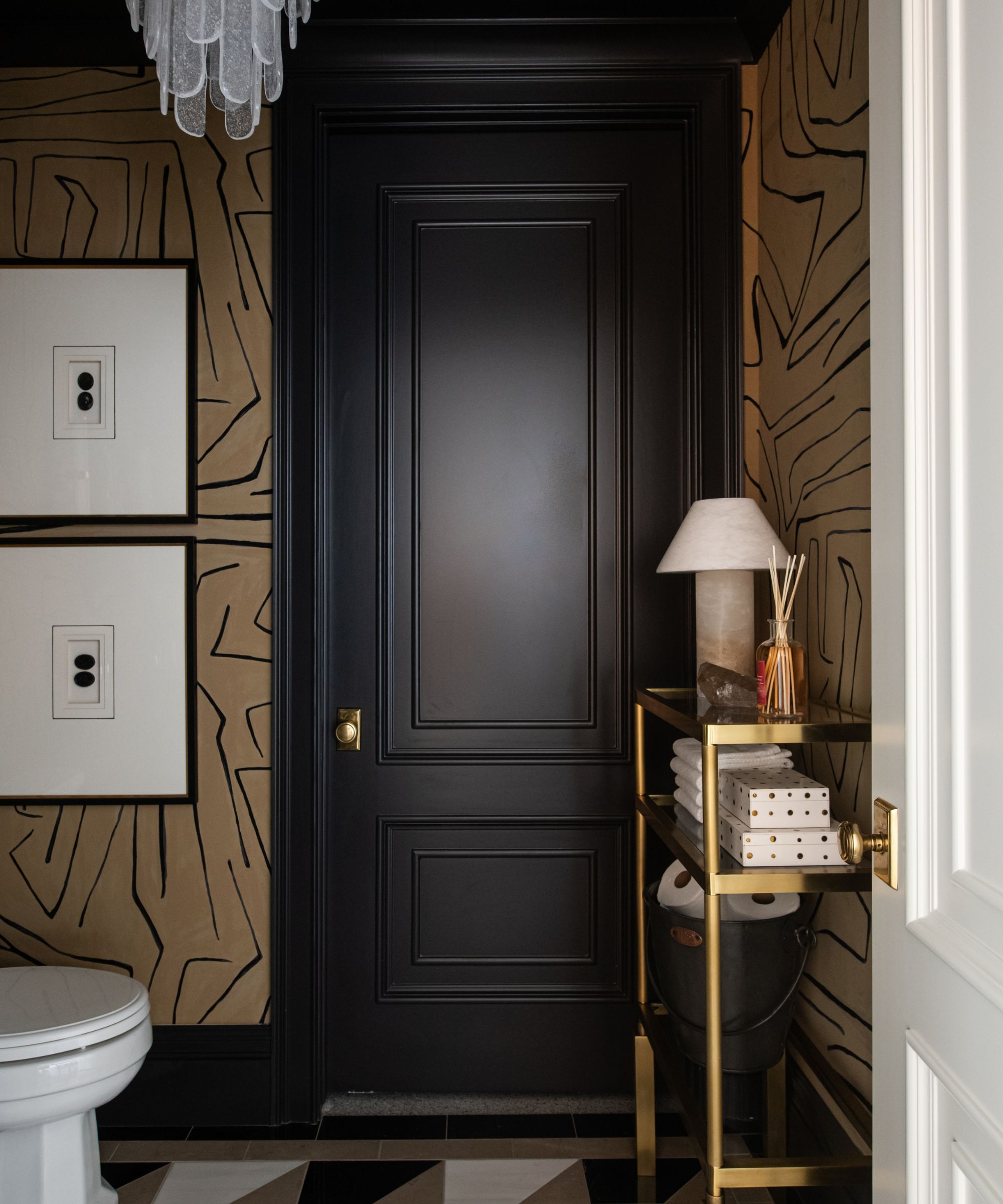 A moody bathroom with black millwork, a gold and black wallpaper, and a gold console decorated with a reed diffuser and lamp