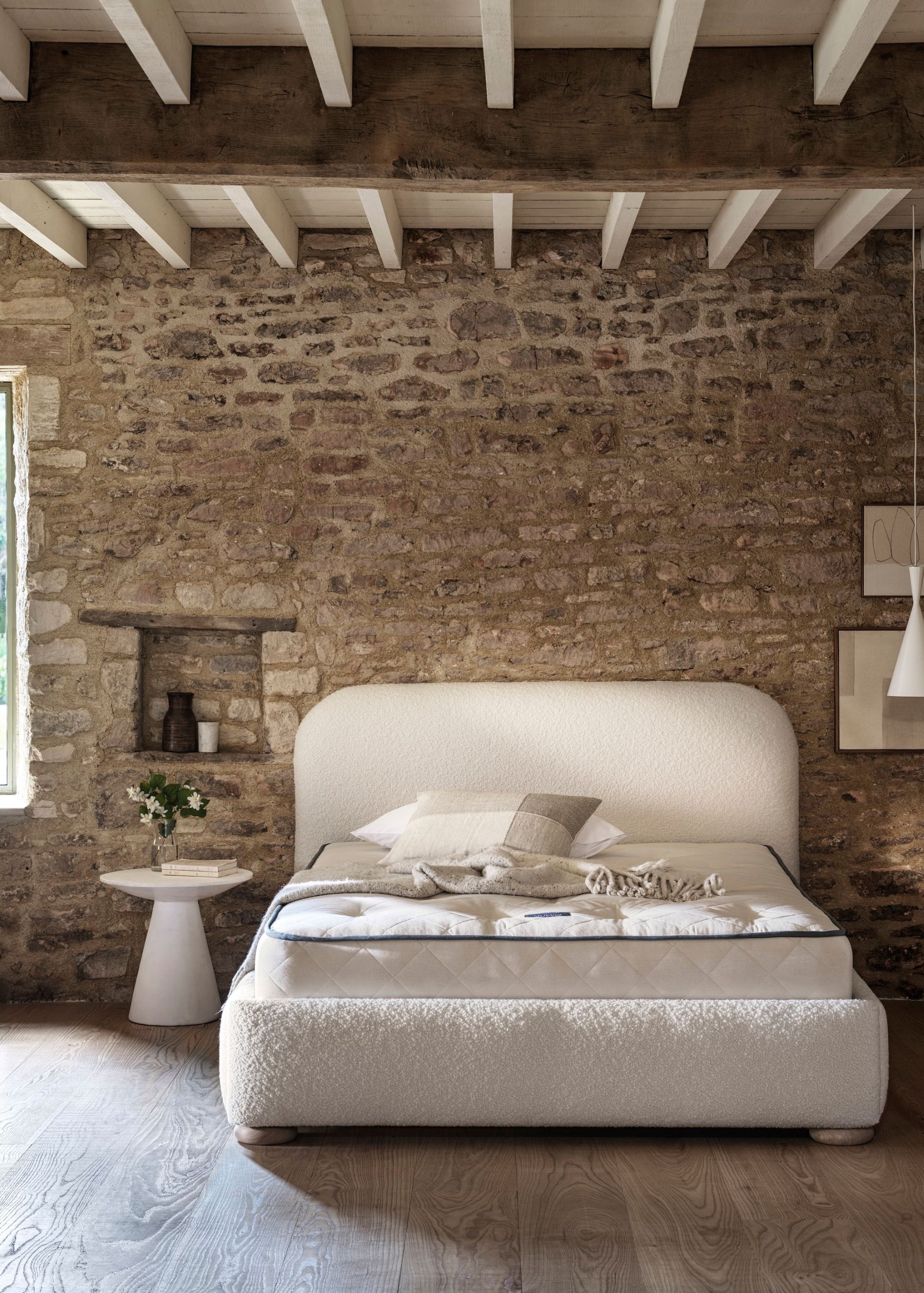 A bare bedroom with a white bed and a natural mattress beside a side table with a vase of white flowers