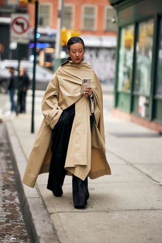 A woman in New York City wearing black wide leg pants, a draped tan trench coat, and black boots