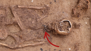 Skeleton lying face-up in reddish soil with a red arrow pointing to a silver amulet at his chin