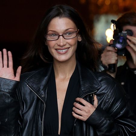 Bella Hadid in Paris for Fashion Week wearing a black '80s leather jacket, a low-cut black top, a brown belt, and pinstripe pants, all by Saint Laurent.