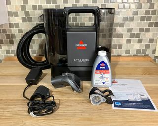 An unboxed Bissell Little Green Cordless Portable Carpet Cleaner, cleaning solution, and instruction manual on wooden worktop with green tiled kitchen wall