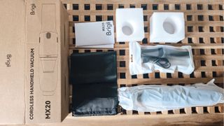 A Brigii handheld vacuum in a white bag, a nozzle in a small white bag, a black drawstring bag, two white foam cubes, a small instruction manual, and a brown cardboard box