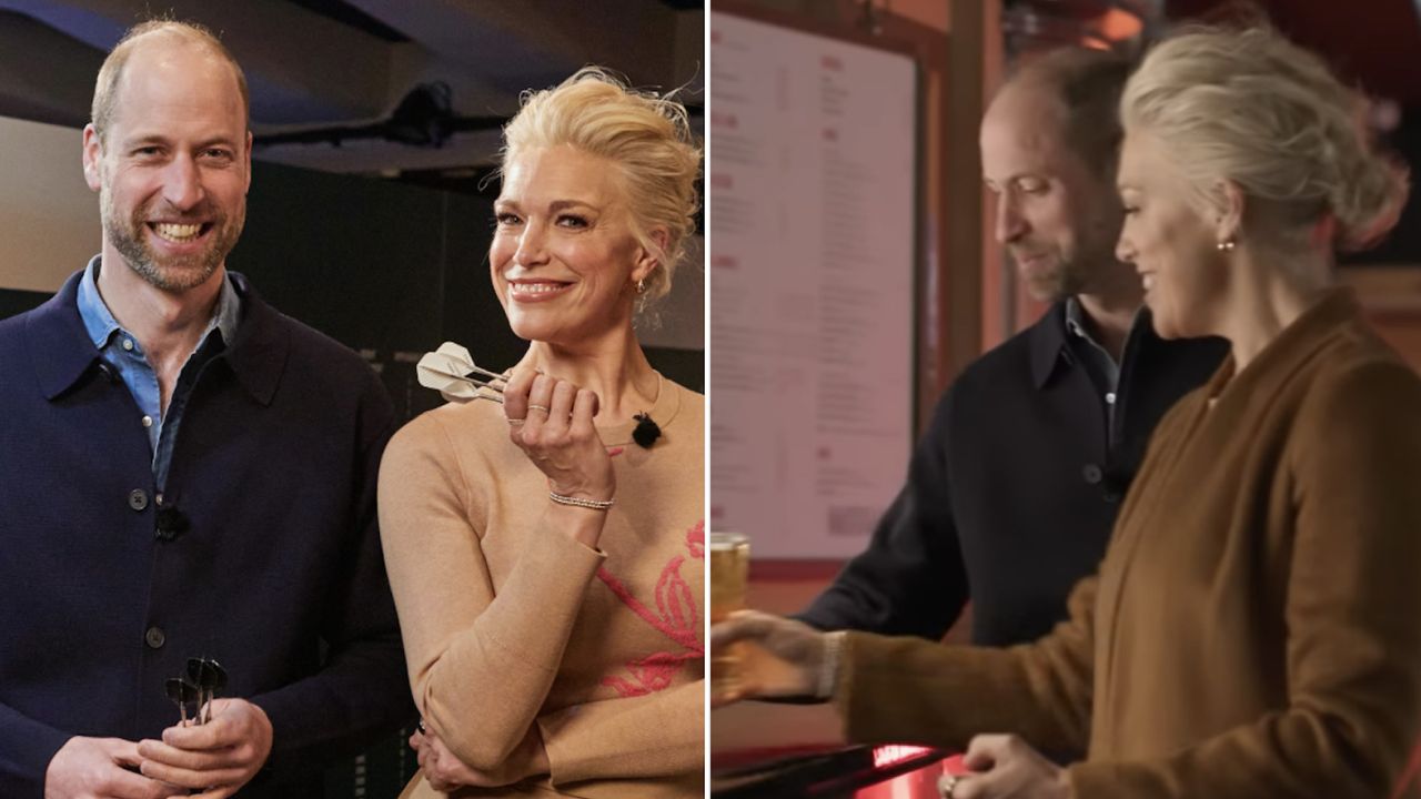 Prince William wears a navy sweater over a blue shirt as he poses Hannah Waddingham who has blonde hair, wears a taupe sweater, and is holding some darts