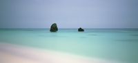 A minimalist, wide-angle seascape showing two dark rocks sitting in the middle of a perfectly still, turquoise ocean under an overcast sky.
