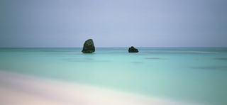 A minimalist, wide-angle seascape showing two dark rocks sitting in the middle of a perfectly still, turquoise ocean under an overcast sky.